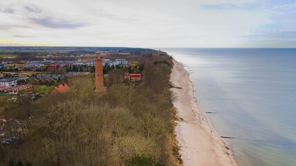 A drone captures Gąski beach, West Pomeranian Voivodeship, Poland, featuring a red brick lighthouse, Baltic Sea, sandy shore, leafless trees on dunes, holiday cottages, hotels, and residences.  © Grzegorz