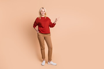 Full length photo of good mood pretty woman wear red sweater pointing finger empty space isolated beige color background