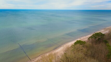 A drone photo showcasing Gąski beach in West Pomeranian Voivodeship, Poland. The image displays the blue-green waters of the Baltic Sea, a wide sandy beach, and leafless trees on the dunes.  © Grzegorz
