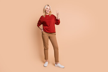 Full length photo of pretty cheerful woman wear red sweater showing v-sign isolated beige color background