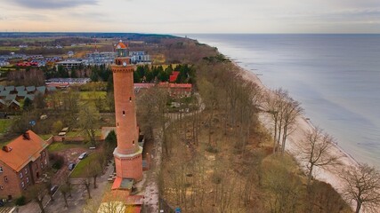 A drone captures Gąski beach, West Pomeranian Voivodeship, Poland, featuring a red brick lighthouse, Baltic Sea, sandy shore, leafless trees on dunes, holiday cottages, hotels, and residences.  © Grzegorz