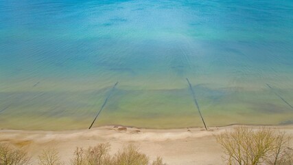 A drone photo showcasing Gąski beach in West Pomeranian Voivodeship, Poland. The image displays the blue-green waters of the Baltic Sea, a wide sandy beach, and leafless trees on the dunes. 