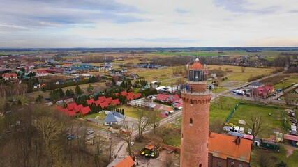 A drone captures Gąski beach, West Pomeranian Voivodeship, Poland, featuring a red brick lighthouse, Baltic Sea, sandy shore, leafless trees on dunes, holiday cottages, hotels, and residences. © Grzegorz