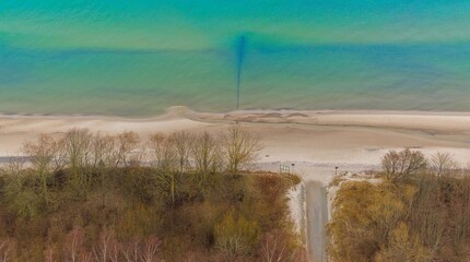 A drone photo showcasing Gąski beach in West Pomeranian Voivodeship, Poland. The image displays the blue-green waters of the Baltic Sea, a wide sandy beach, and leafless trees on the dunes.  © Grzegorz