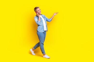 Fototapeta na wymiar Full body photo of attractive young woman walk talk mobile phone dressed stylish denim clothes isolated on yellow color background