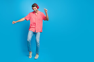 Full size photo of optimistic man dressed striped shirt in headphones dancing near empty space offer isolated on blue color background