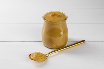 Spoon and glass jar of tasty mustard sauce on white wooden table
