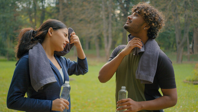 Tired exhausted Indian man and woman relaxing after sports in park outdoor Arabian fitness couple in city sport people wipe sweat with towels exhausted relax pause break drinking bottled water refresh