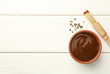 Tasty barbeque sauce in bowl, brush and peppercorns on white wooden table, top view. Space for text