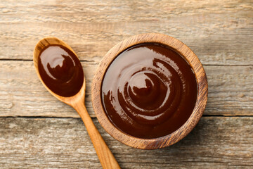 Tasty barbeque sauce in bowl and spoon on wooden table, top view