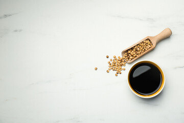 Soy sauce in bowl and scoop with soybeans on white marble table, flat lay. Space for text