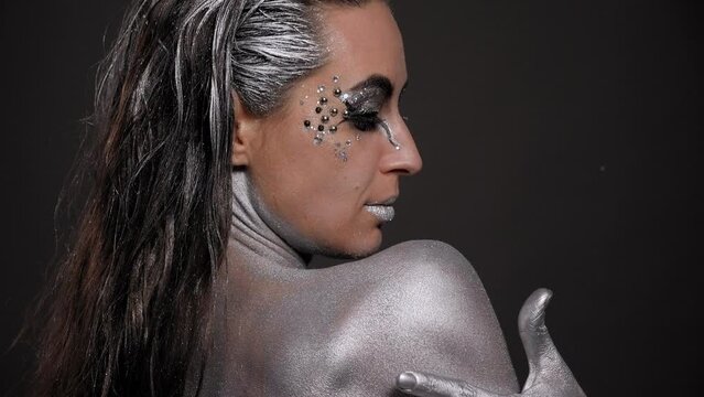 Close-up of a gorgeous female model with silver skin and hands, with bright makeup and stones, she poses in the studio on a black background. Fashion portrait of a young woman.