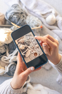 A woman takes pictures of yarn, threads and a cup of coffee on her smartphone.