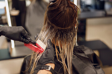 Close up of hairdresser is dying hair of client with brush while working in professional salon