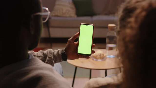Dolly-out back-view shot of interracial couple sitting on couch in cozy living room and looking at smartphone with green chroma key screen