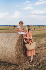 Mom and little daughter in a field of harvested wheat against the blue sky. Mother and daughter look into each other's eyes. Mom is holding a wicker linen bag in her hands.