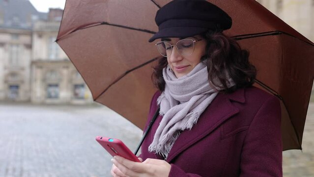 Young adult woman holding an umbrella using mobile phone in a rainy day at city street. Technology concept.