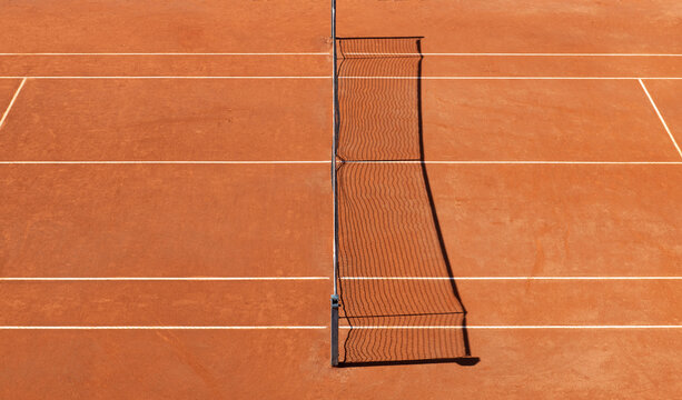 A top view of an empty clay tennis court with tennis net, lines, and orange surface
