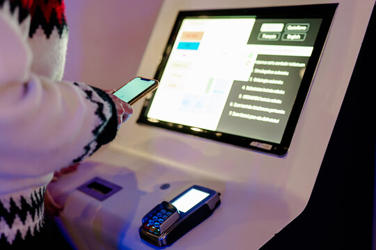 Anonymous person using a smartphone to interact with a digital kiosk touch screen display next to a card payment terminal