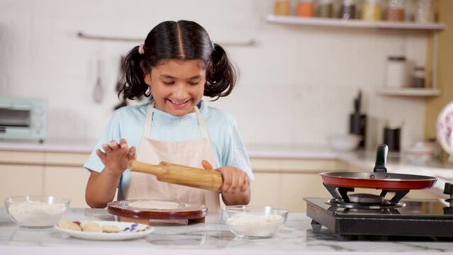 Happy Girl Kid looking at camera by showing chapati while doing chapati at kitchen - concept of childhood fun, playful activities and hobbies