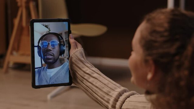 Over shoulder of Caucasian girl video calling her African American boyfriend through digital tablet while at home, having long distance relationship