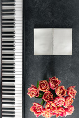 Piano, bouquet of tulip flowers and notepad on a dark background, top view.