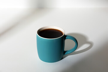A cup of coffee stands on a white background with a shadow from the window in the morning light