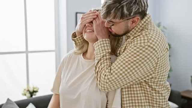 A man surprises a woman with a blindfold game in a cozy living room, expressing love and joy.