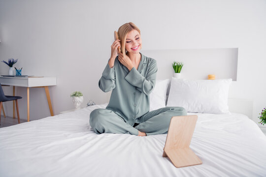 Photo of good morning concept beautiful blonde girl brushing her hair with comb looking at mirror sitting on bed indoors light interior