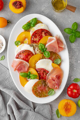 Salad of sliced multi-colored tomatoes, fresh peaches, mozzarella, prosciutto and basil on a gray concrete background. Delicious summer salad.