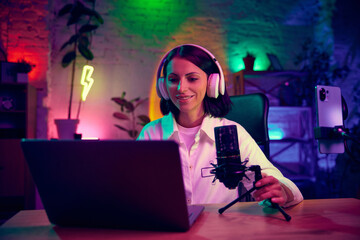 Obraz na płótnie Canvas Young woman, blogger, streamer in headphones, sitting at home in modern room and leading online stream, podcast. Blogging, online services, streaming, education concept