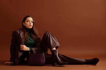 Fashionable confident woman wearing faux leather suit blazer, pants, pointed toe ankle boots, with trendy purple bag, posing on brown background. Full-length studio fashion portrait. Copy, empty space