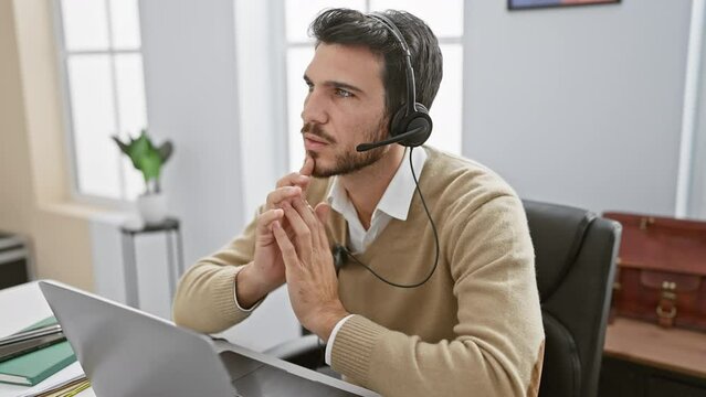 Handsome young hispanic man with a beard working in an office, wearing a headset and concentrating on a laptop screen.