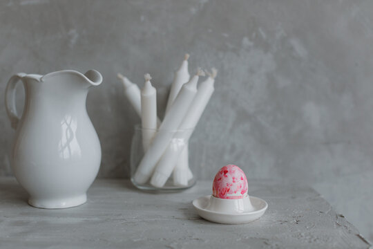A painted Easter egg on a stand, candles in a glass vase, a jug on the mantelpiece. Minimalistic still life in gray.