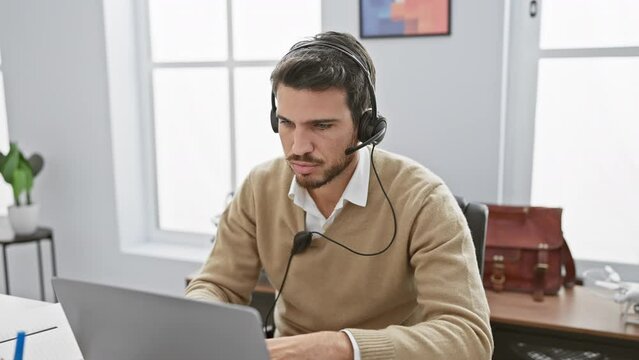 Young bearded man with headphones at a laptop in a modern office, feeling tired and closing his computer.