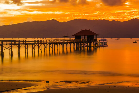 A wooden pier stretches out into a calm bay at sunset, with mountains in the background.