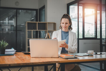 Business analytics concept, A smiling young professional woman using a laptop and holding a...