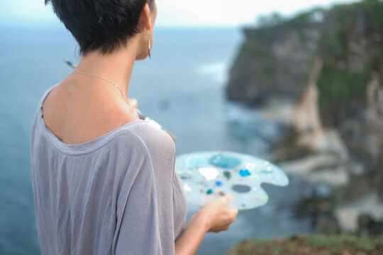 Portrait of a middle-aged woman artist who stands on a hill and mixes blue shades of paint with a brush on a palette, looking at the ocean. The artist paints a landscape painting in outdoors.