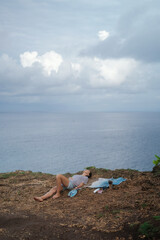 A woman artist lies on the edge of a cliff next to a painting she painted with acrylic paints on canvas. Nature inspiration concept by the ocean. Drawing outdoors.