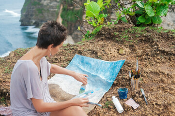 From the back, a woman artist sits on a rock by the ocean and paints a landscape from life. Sea...
