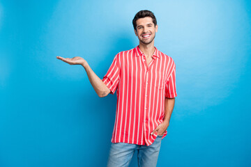Photo of cheerful funky man dressed striped shirt holding arm empty space isolated blue color...