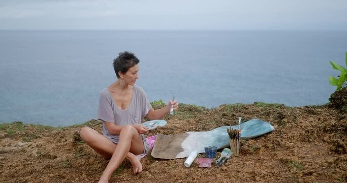 A woman artist sits on a cliff by the ocean and paints a landscape painting from life. Sea painting with acrylic paints on canvas. Next to picture is a palette of paints, a plein air in the open air.