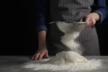 Making bread. Woman sifting flour over dough at table on dark background, closeup