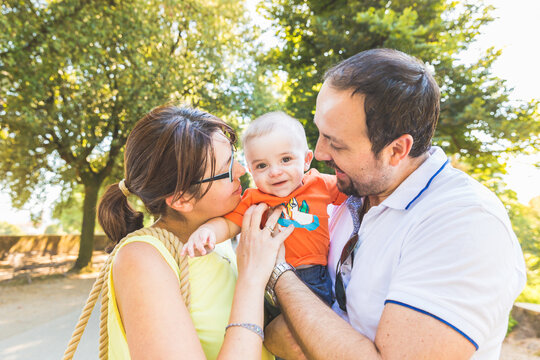 Happy parents kissing their baby with affection during a family outing - Joyful family enjoying a sunny day in the park