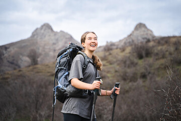 Smiling young traveler woman wearing hiking clothes holding trekking poles, traveler looking up and...