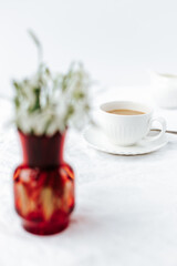 in the foreground out of focus there is a red vase with snowdrops, in the background there is a white cup of coffee in focus