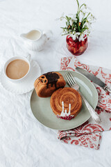There is a two round croissant on a beige plate.Next to it are a napkin with red patterns and vintage cutlery,a cup of coffee, a milk jug and a red vase with snowdrops.White backgrounds