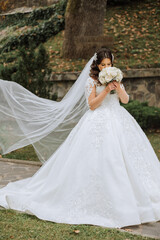 A brunette bride in a voluminous white dress holds a bouquet, poses. The veil is thrown into the air. Beautiful hair and makeup. Wedding photo session in nature.