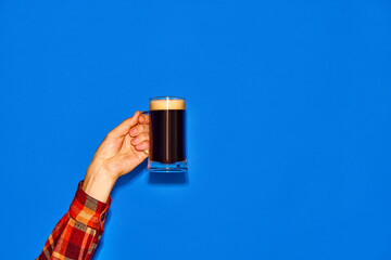 Male hand holding mug with dark, foamy beer isolated on blue background. Traditional taste. Concept...