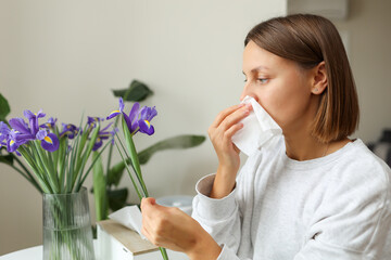 Allergic young woman holds iris flower, covers nose with paper tissue has runny nose, sneezes from...